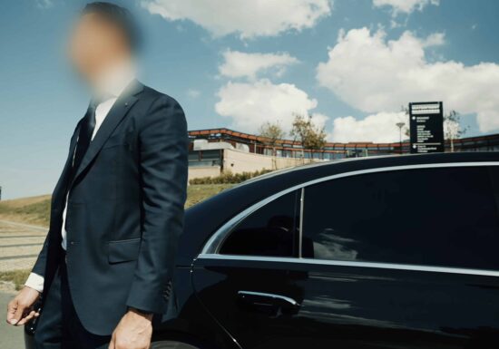 How to hire a bodyguard | A businessman in a suit standing next to a black luxury car under a clear blue sky.