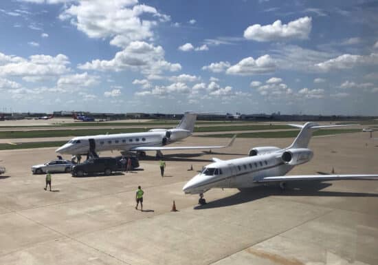 International bodyguard services and executive protection | Two private jets parked on an airport tarmac under a bright blue sky with scattered clouds. Ground crew members in yellow vests and other personnel are seen near the planes, along with several vehicles, including a black SUV and a white car. In the background, several commercial airplanes are visible on the runway.