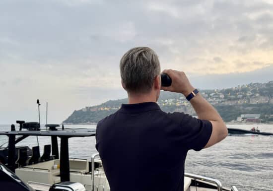 Yacht Event Security Services | Man using binoculars on a yacht overlooking the sea and hilly coastline.
