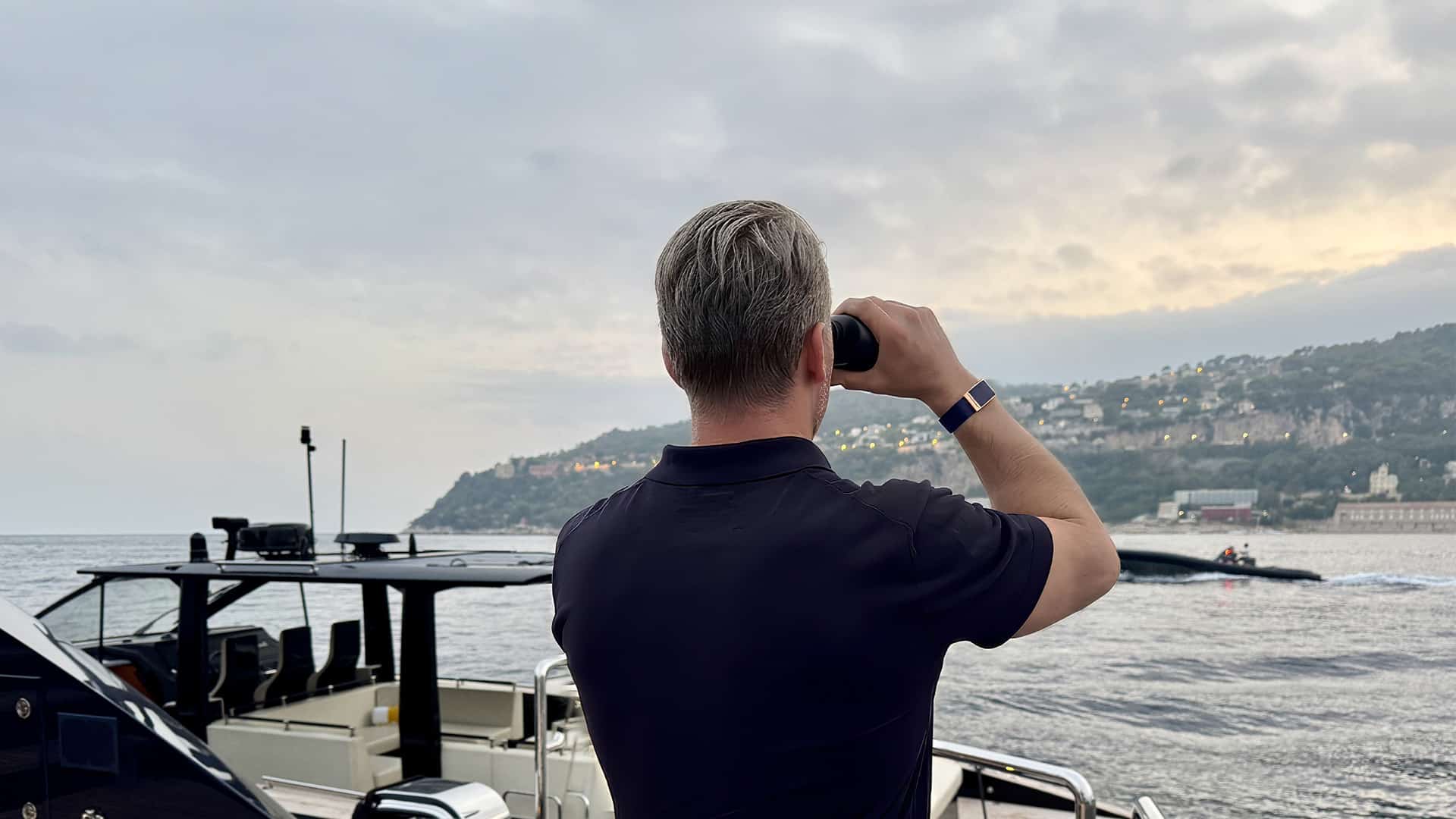 Yacht Event Security Services | Man using binoculars on a yacht overlooking the sea and hilly coastline.