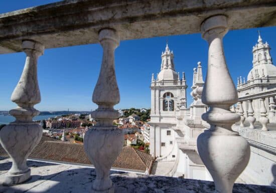 Bodyguard services Portugal. A scenic view of Lisbon, Portugal, captured through the stone balustrade of a cathedral, showcasing the city's rooftops and the Tagus River.