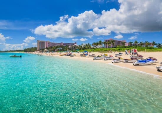 Bodyguard services on the Bahamas. A vibrant beach scene in the Bahamas with clear turquoise water, jet skis lined up on the shore, and a backdrop of hotels and palm trees.