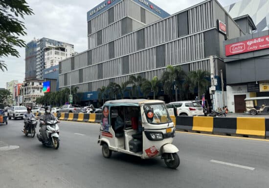 Bodyguard services in Cambodia. A bustling street in Cambodia featuring motorbikes, a tuk-tuk, and modern buildings including a bank.