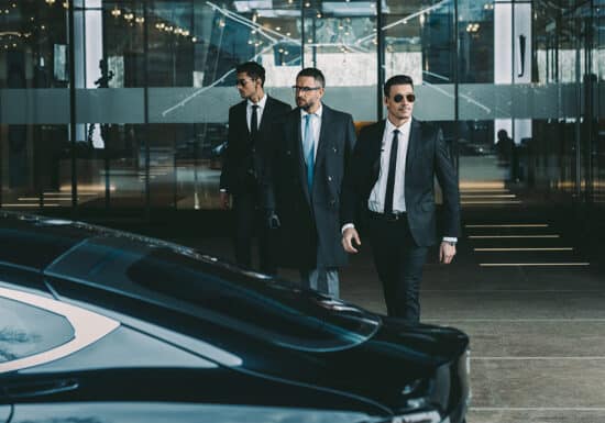 How to Hire a Bodyguard. Three security men in suits and sunglasses walking purposefully beside a black luxury car.