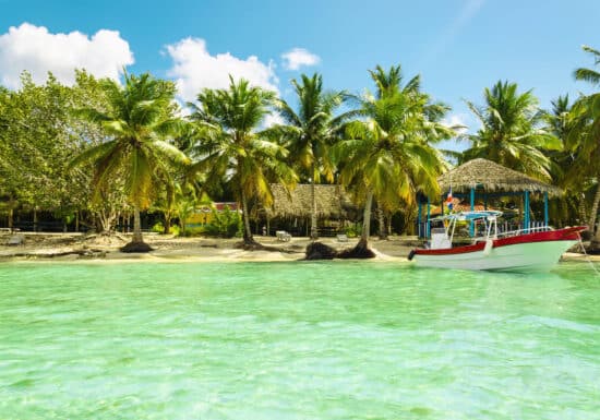 Bodyguard Services in the Caribbean. A tropical beach scene featuring a boat anchored in clear turquoise water, with palm trees and thatched huts along the sandy shore under a blue sky with scattered clouds.