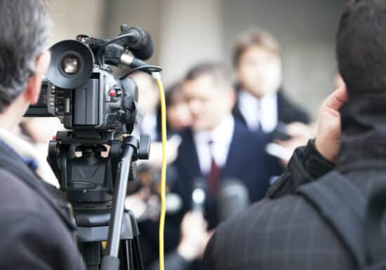 Media Backwatch Service. Close-up of a video camera recording a press conference with journalists and a spokesperson in the background.