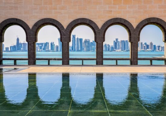 Bodyguard services in Qatar. View of a modern city skyline through arched windows from a serene waterfront terrace, with reflections on the still water.