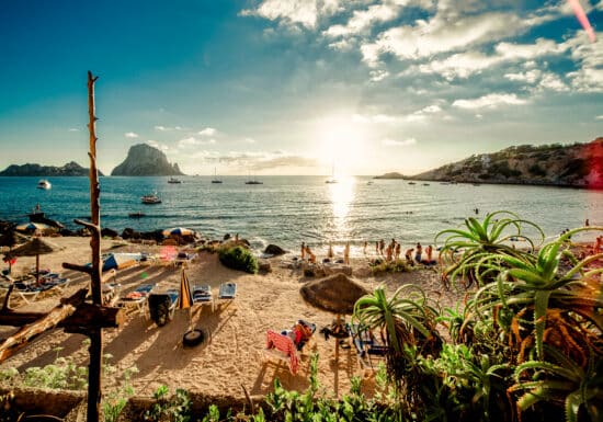 Bodyguard Services on Ibiza. A picturesque beach with people relaxing under umbrellas, surrounded by a stunning coastal landscape with boats in the water.