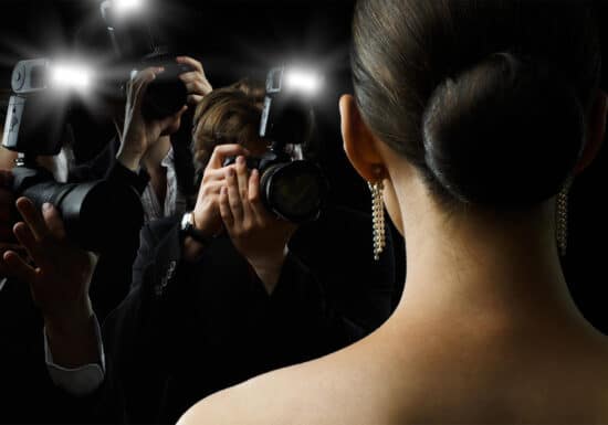 Celebrity Bodyguard & VIP Protection Services. A woman with her hair in a bun and wearing pearl earrings faces a crowd of photographers taking her picture, their camera flashes illuminating the dark background.