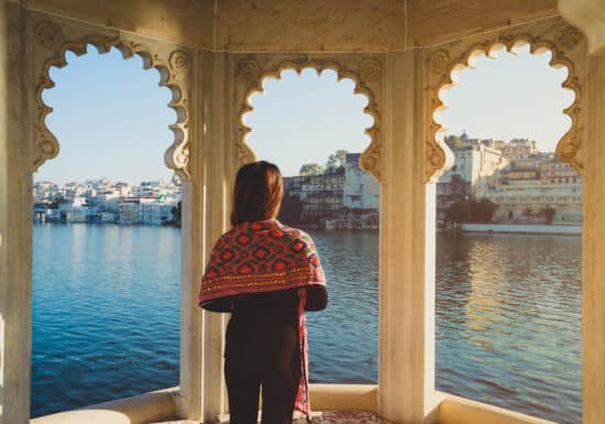 Solo Travel in India. A woman stands on a balcony overlooking a lake, with city buildings in the background, framed by ornate arches.