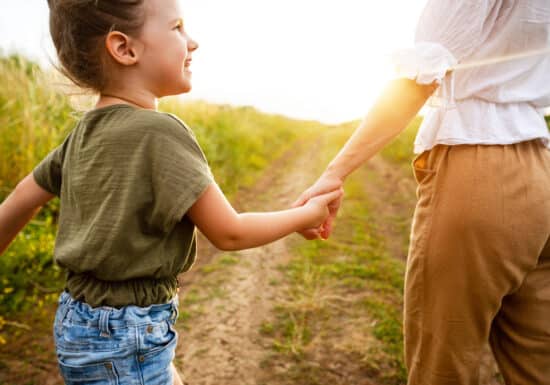 Hiring a bodyguard for your child. A child holding an adult’s hand, walking down a sunlit path in a grassy field.