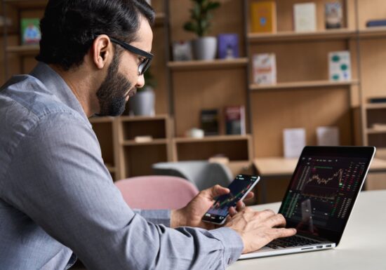 Bodyguards for Crypto Millionaires. A man using a smartphone and laptop to monitor stock market data.