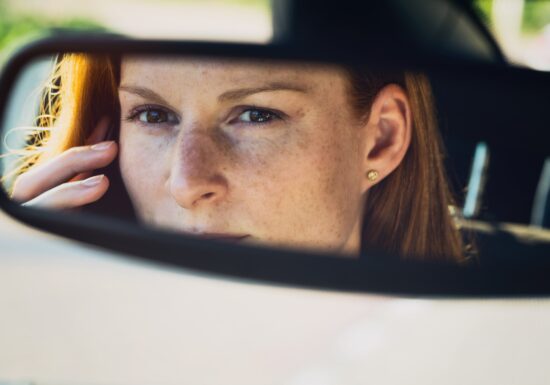 Female Bodyguards. A woman’s face reflected in a car’s rearview mirror, showing her serious expression and red hair. She is holding a phone in her hand.