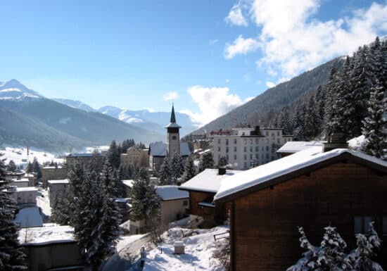 Secure transportation in Davos. Snowy village with mountains, church steeple, and evergreen trees under a clear sky.