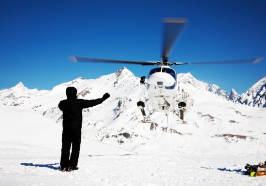 Ski Holiday Bodyguard Service. A person dressed in black winter gear stands on a snowy mountain slope, directing a white helicopter landing in front of them. Snow-covered peaks and a clear blue sky form the backdrop.