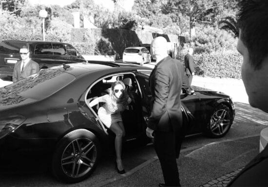 How Much Does It Cost to Hire a Bodyguard. A woman in sunglasses stepping out of a black luxury car, surrounded by security personnel.