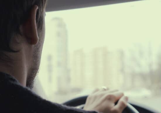 Intelligence Gathering. Person driving a car, seen from the backseat. A dashcam is mounted on the windshield and buildings and other cars are visible outside.